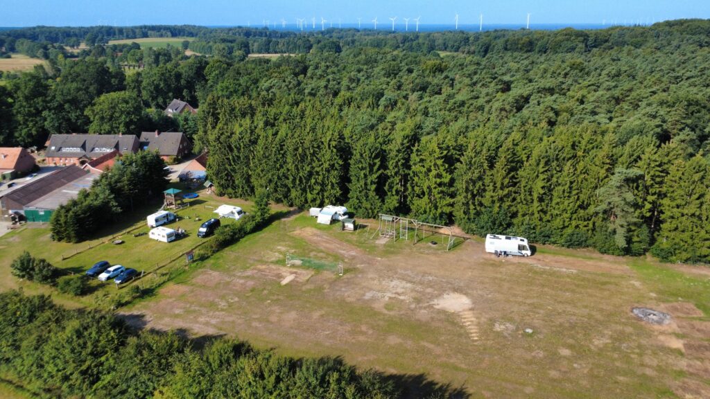 Luftaufnahme eines ruhigen Stellplatzes am Waldrand. Mehrere Wohnmobile und Wohnwagen stehen auf einer Wiese nahe dem Ferienhofs. Im Hintergrund dichter Wald, im Vordergrund ein Spielplatz und weite Grünfläche.